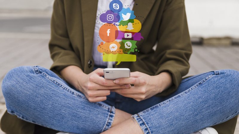 Person sitting cross legged on mobile phone with social media icons above